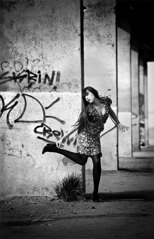 Frau im gemusterten Kleid und schwarzen Strumpfhosen posiert dynamisch an einer Graffiti-Wand